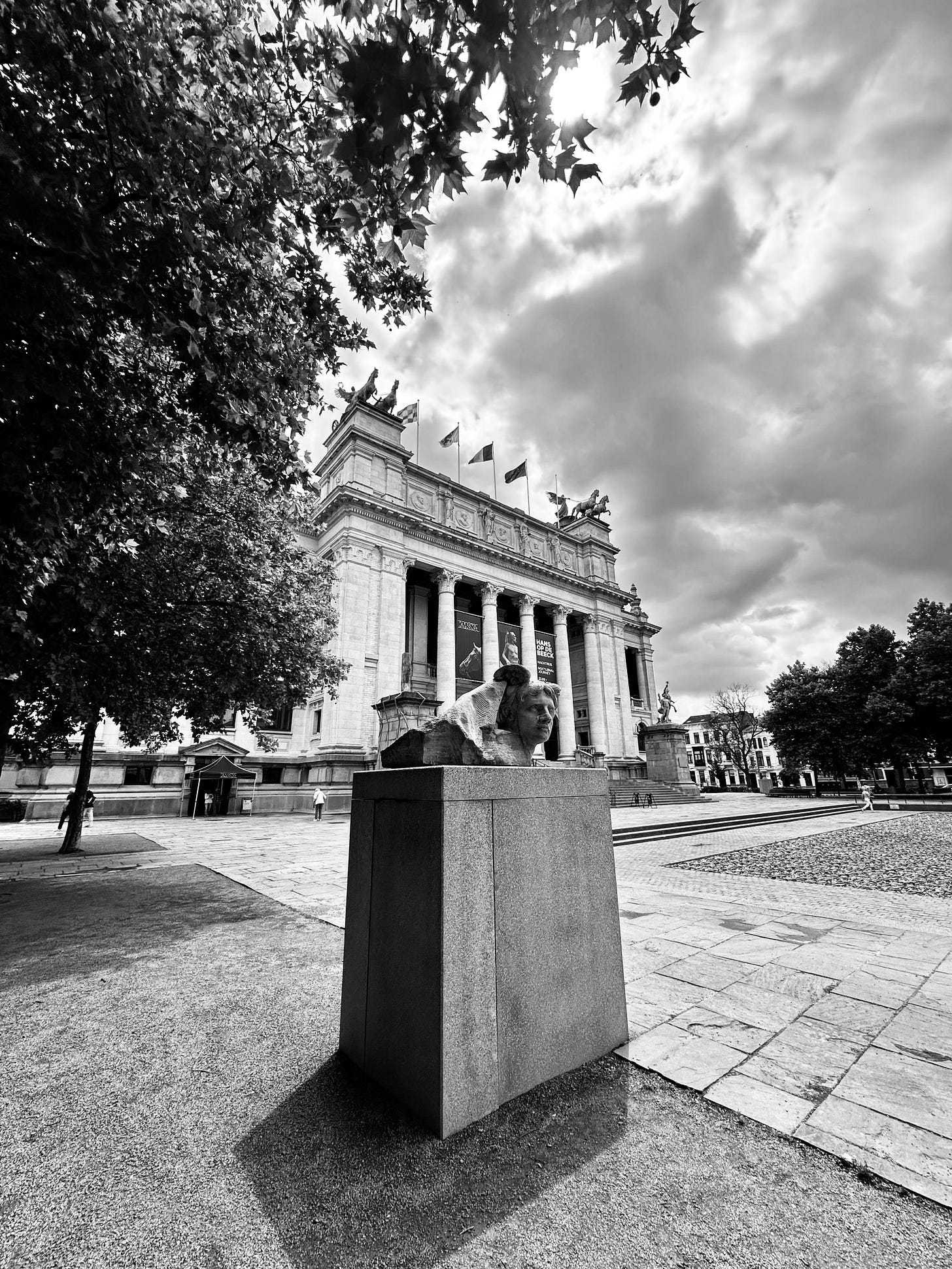 Black and white photo that I took in the summer of 2025 of the newly reopened Koninklijk Museum voor Schone Kunsten in Antwerp. It is built in a neoclassical style with imposing columns and decorative carvings on its facades. In the the foreground is a rectangular plinth with a contemporary sculpture of a bust.