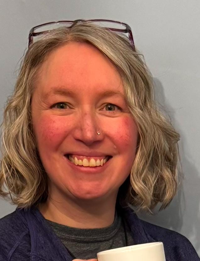A headshot of Marion McLaughlin. They have grey, shoulder-length hair and glasses perched on top of their head. They are white and smiling and have a nose stud.