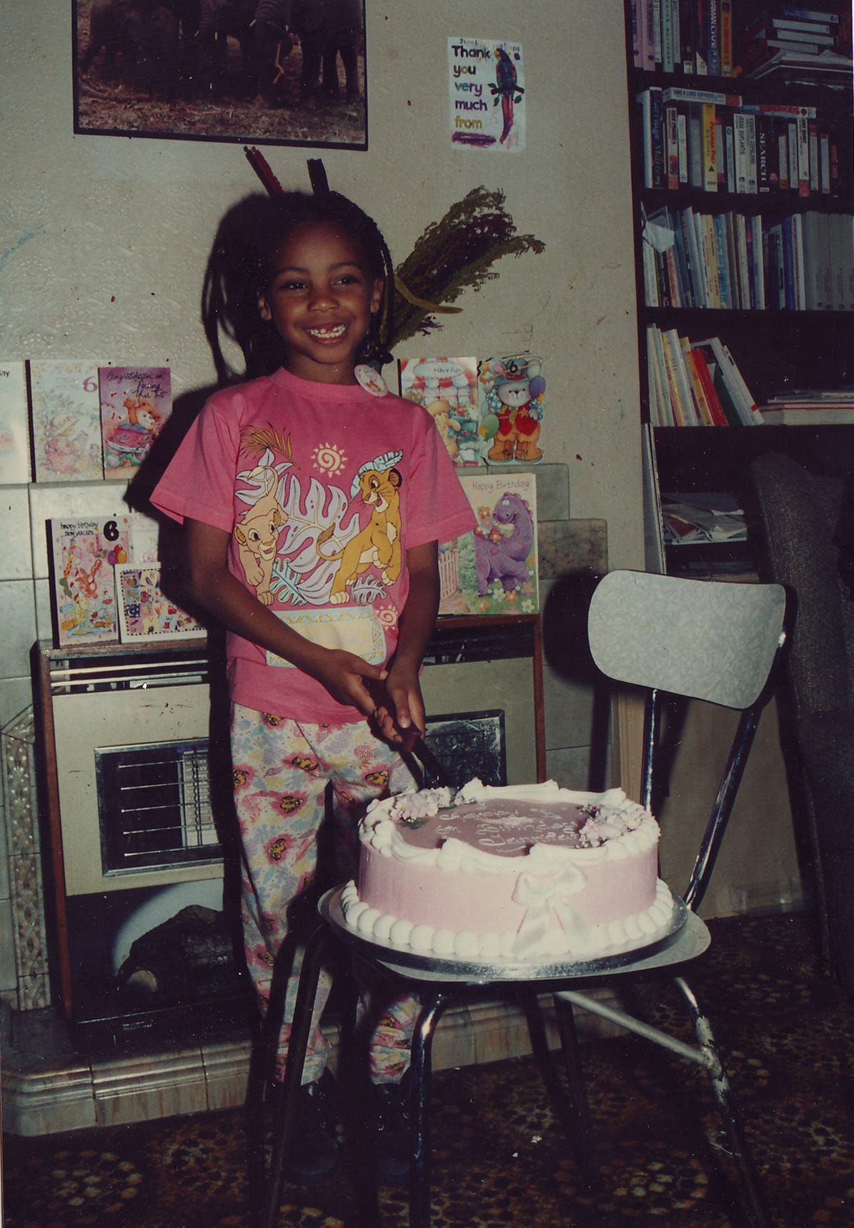 Young girl wearing a pink Lion King t-shirt and matching leggings cuts a large pink cake balanced on a chair. She stands in a very 90s living room, with an electric fireplace behind her with birthday cards.