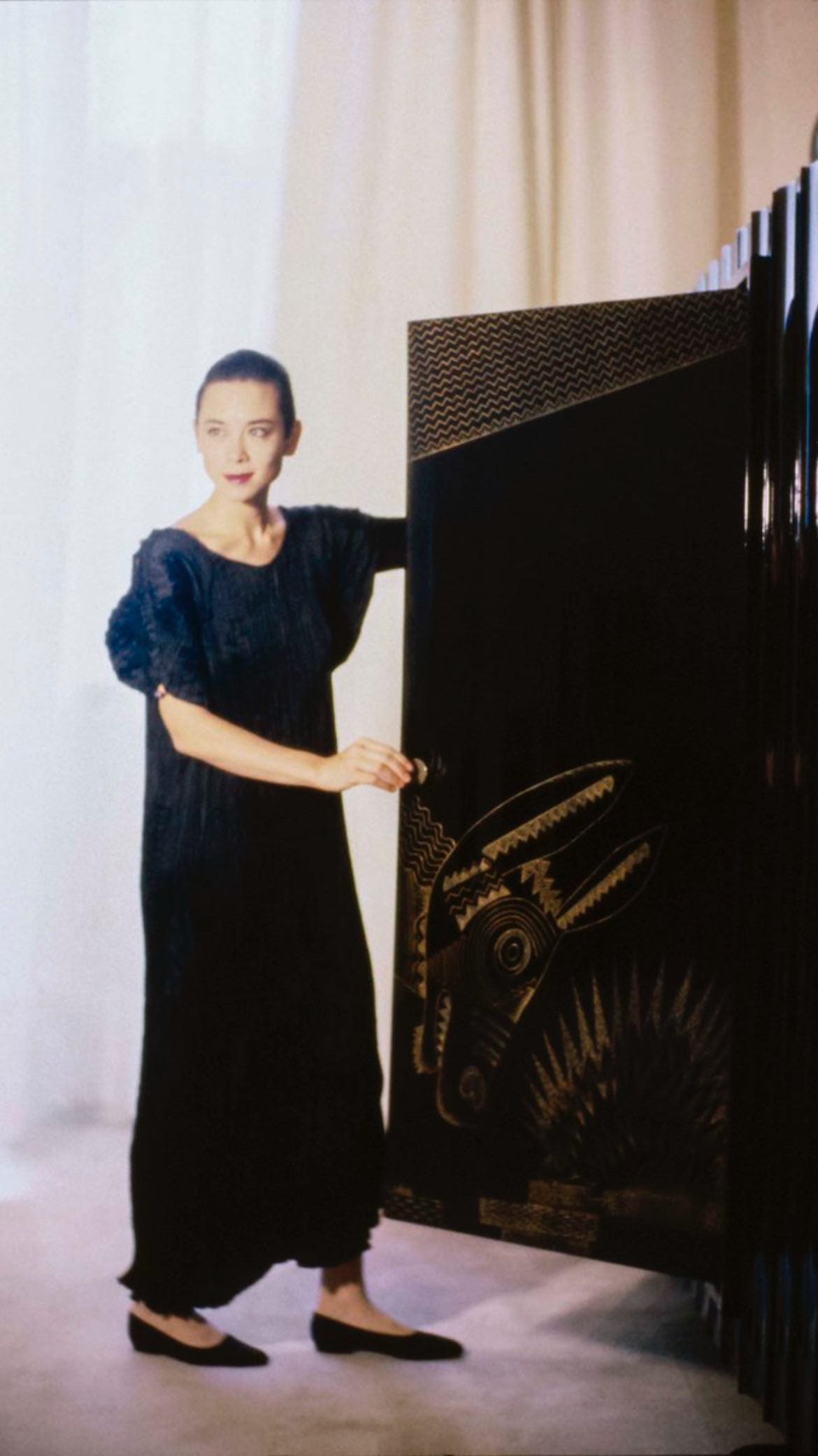 A short-haired woman wearing a loose, tea-length black dress and flats while opening a vintage lacquered wardrobe from the Art Deco period.