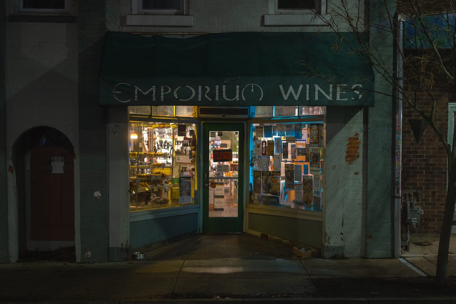 The dimly lit storefront of Emporium Wines, showcasing bottles and various posters through the large glass windows, with a “Closed” sign hanging at the door.
