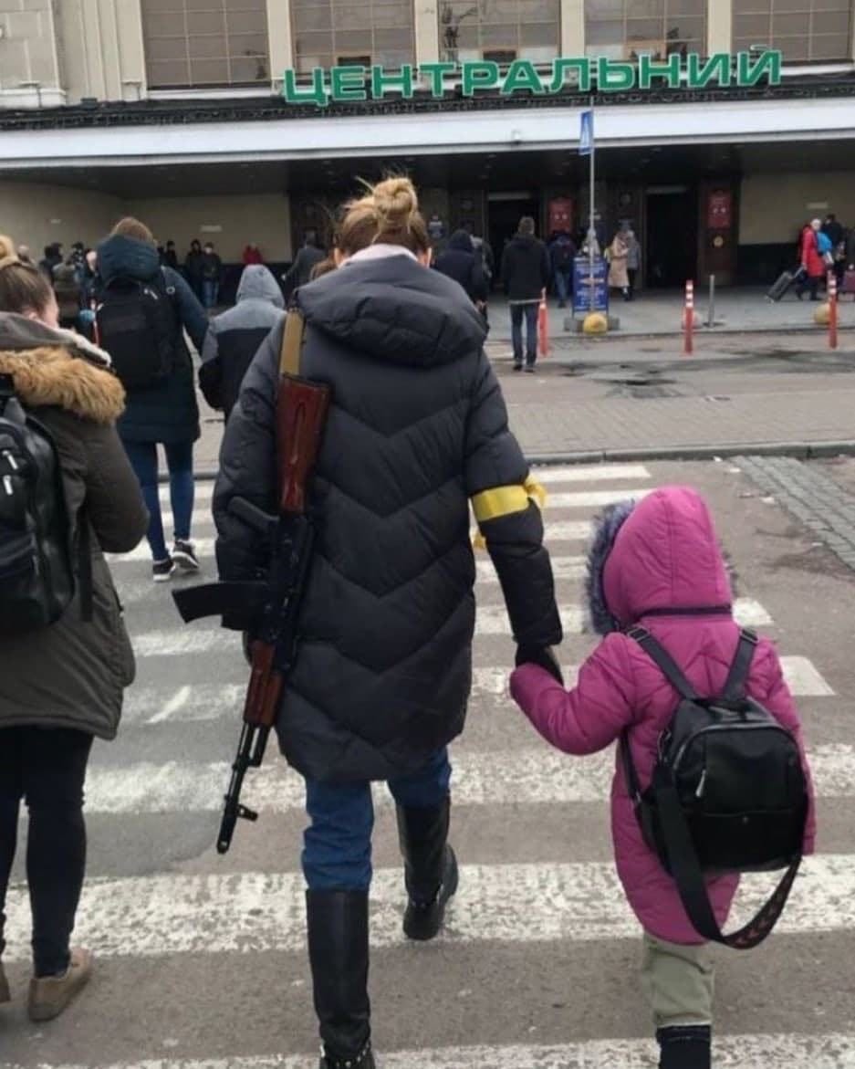 A woman walks a child toward the entrance to a building. They are holding hands. The woman -- in a black coat and yellow armband -- has a military-style rifle slung over her left shoulder. The child -- in a purple coat -- has a backpack slung over their shoulders.