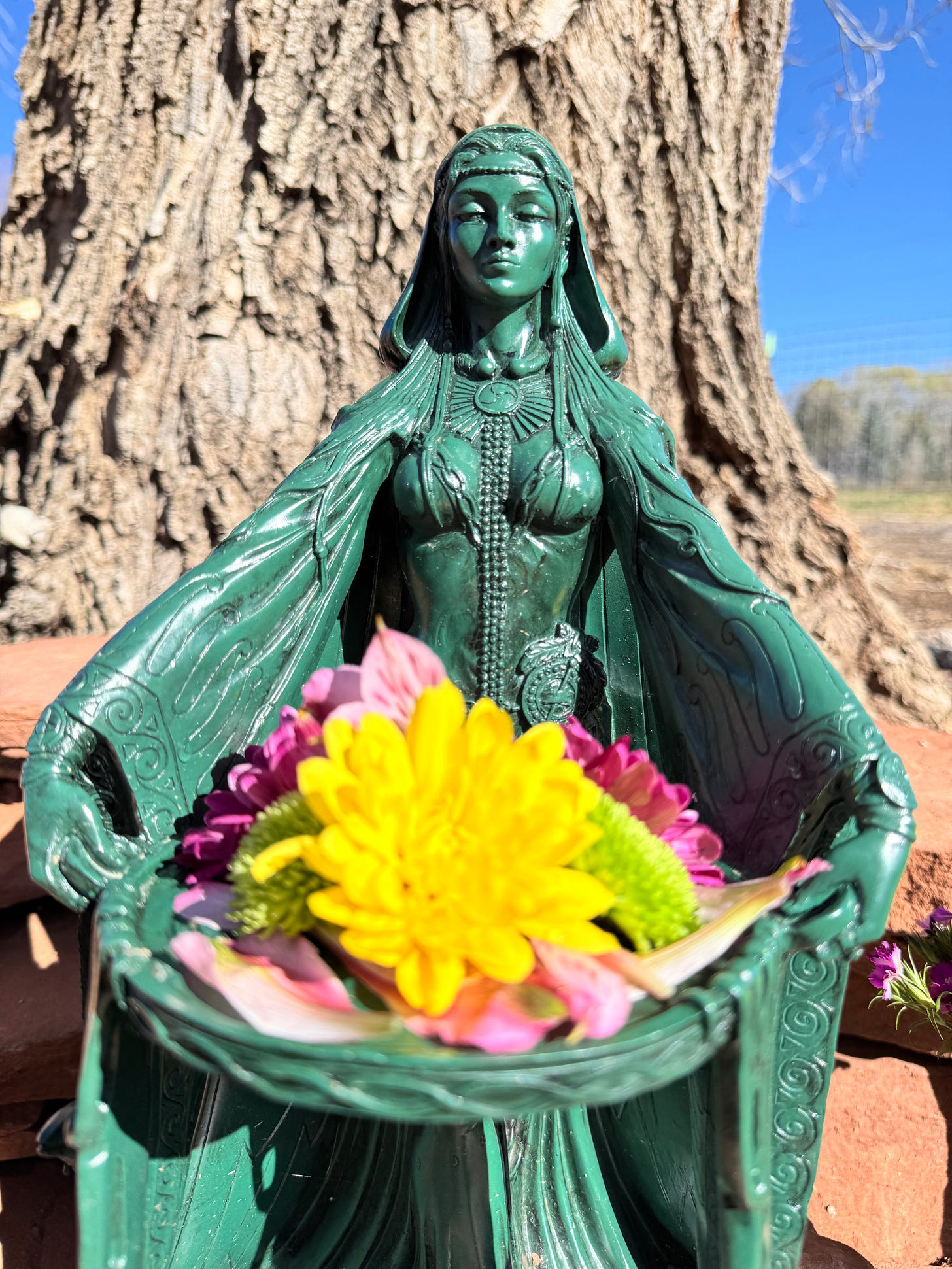 Ostara altar offering, green goddess statue holding bright spring flowers against sunlit tree bark and blue sky