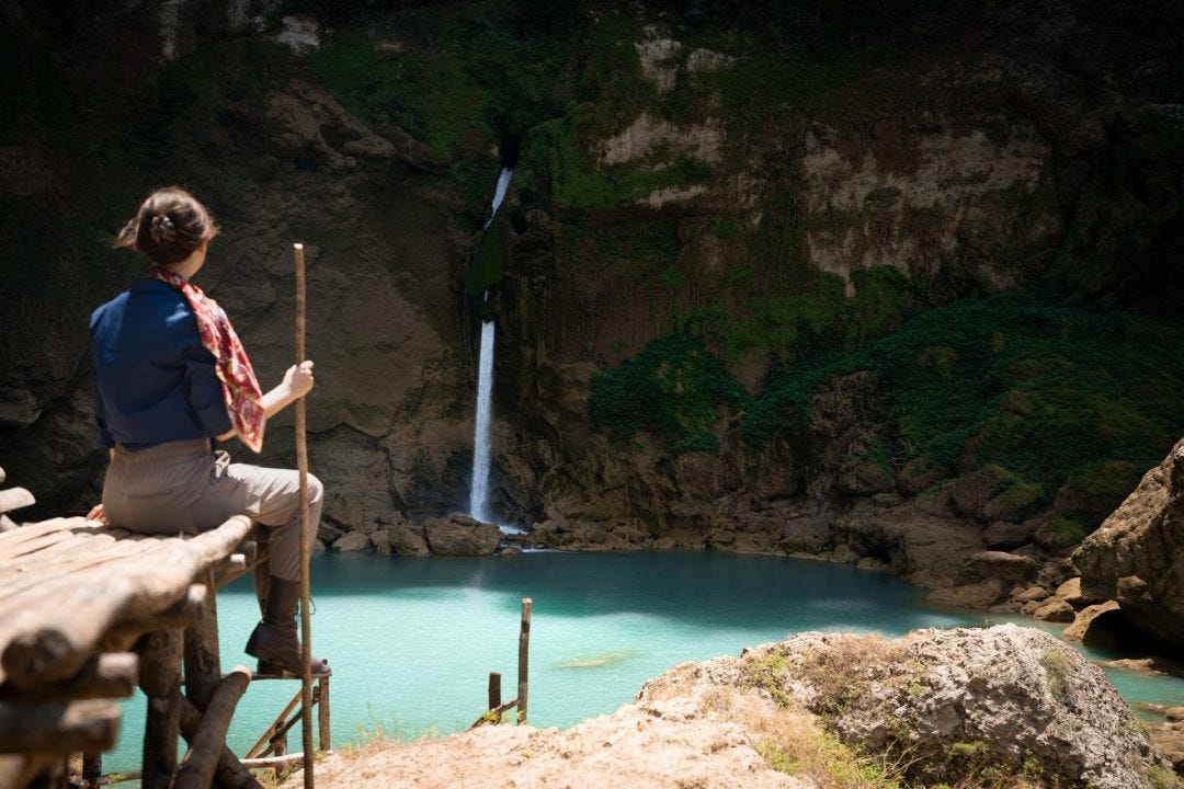 Travel Blogger Jordan Gassner looking out toward Matayangu Waterfall in an explorer outfit and holding a walking stick in Sumba Indonesia