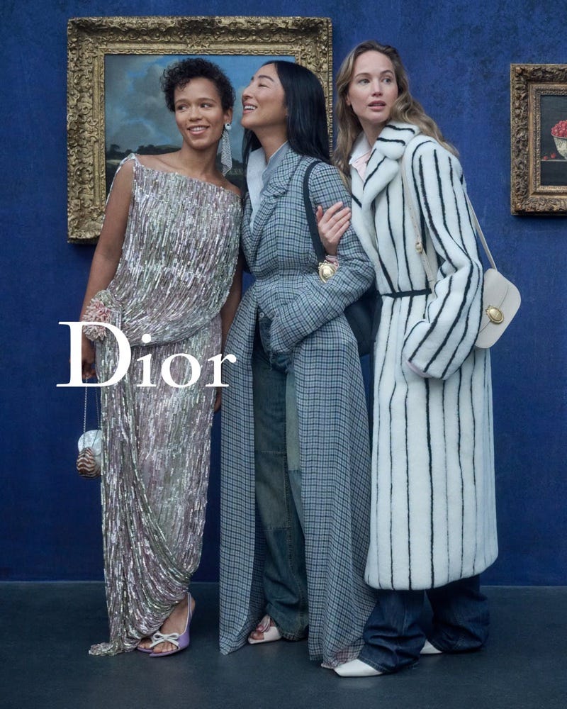 Three women in Dior, in a sequined gown, a grey coat and a striped white coat, against a blue gallery wall hung with gilt-framed paintings