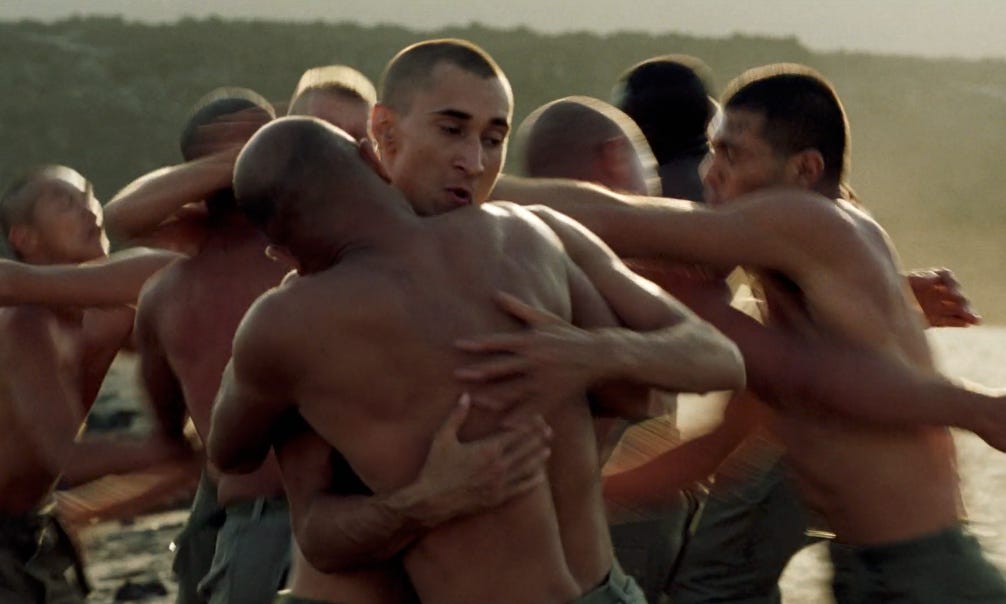 Shirtless soldiers, centered on Grégoire Colin, perform grappling exercises on the beach in a still from Claire Denis's Beau Travail.