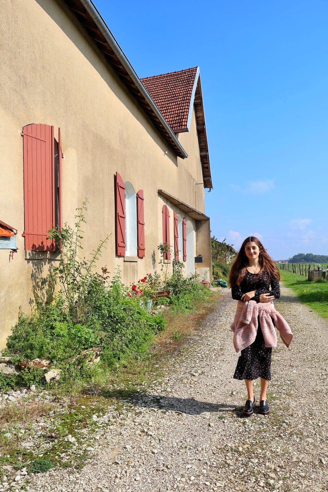 Jeune femme marchant le long d’une maison beige aux volets rouges, sous un ciel bleu éclatant. Jeune femme marchant le long d’une maison beige aux volets rouges, sous un ciel bleu éclatant.
