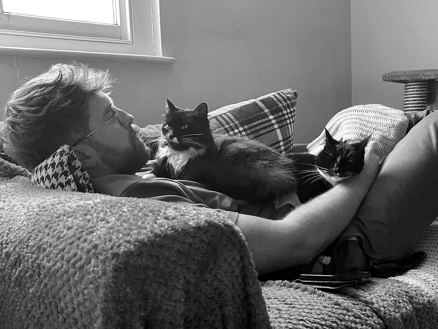 Daniel Kelly, a writer based in Somerset, is laying on a small chair with his two black cats sat on top of him, while playing video games.