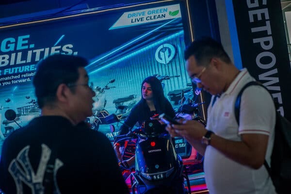 A woman sits on an electric motorcycle in front of a marketing poster that says “Drive Green Live Clean.”