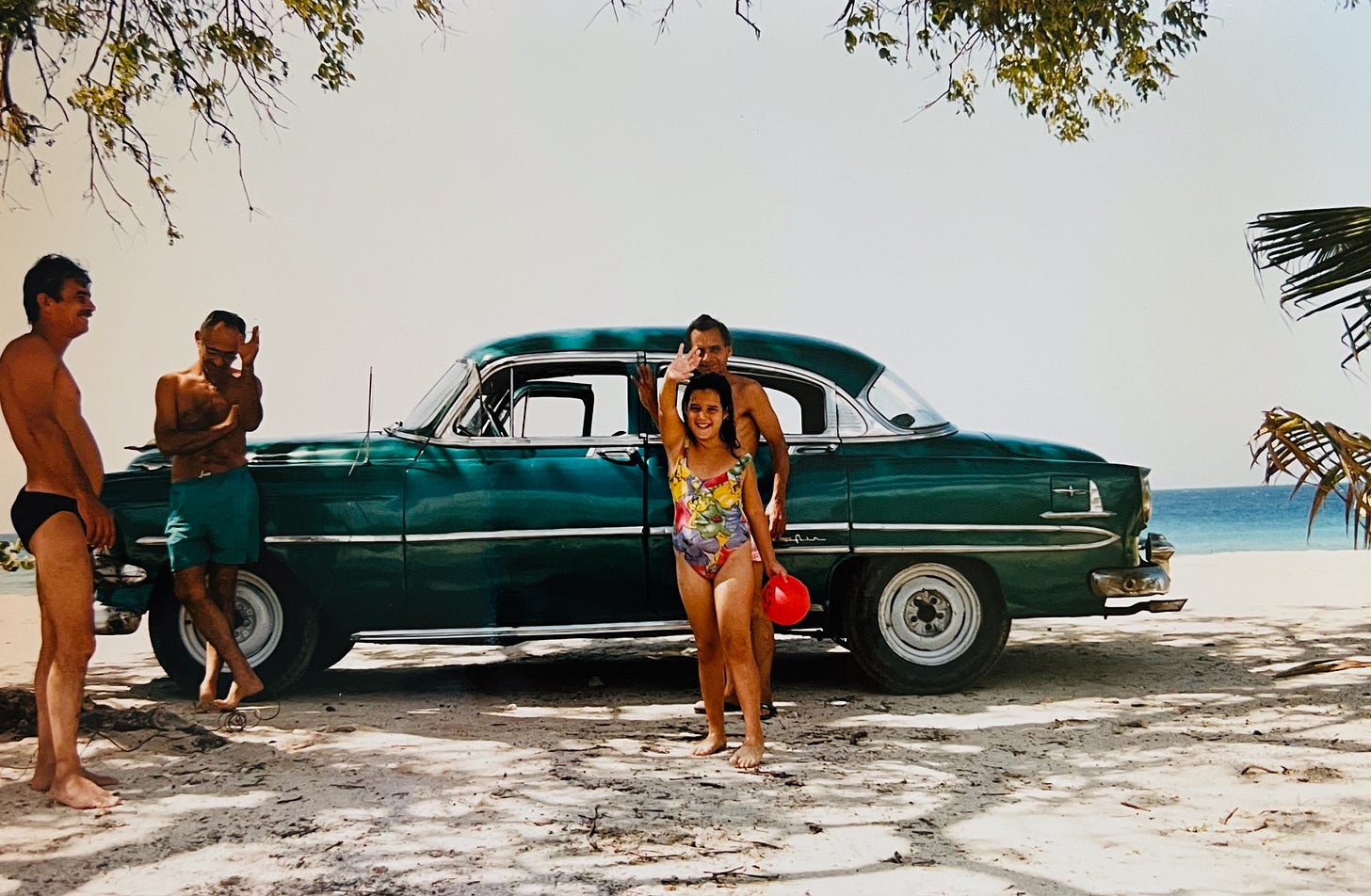 1950s-style car in green on tropical beach with family, including young girl waving to the camera and smiling.