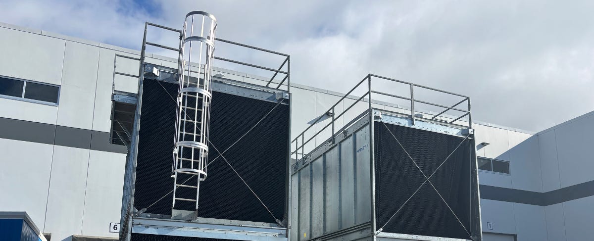 Two large industrial cooling units with protective railings and a ladder are positioned outside a building. Two large industrial cooling units with protective railings and a ladder are positioned outside a building.