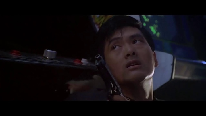 Chow Yun-Fat hides behind an arcade machine while holding a pistol, looking up cautiously during a shootout.