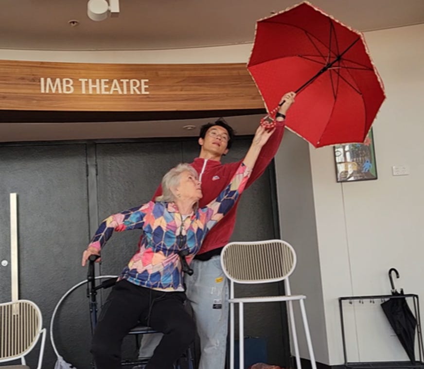 Susan Kennedy and Bubblez in front of a door labeled 'IMB THEATRE'. Susan is seated in a walker, reaching towards a red umbrella that Bubblez is holding. Susan is wearing a bright, multi-coloured patterned top and black pants. Bubblez is wearing a red jacket and light coloured pants.