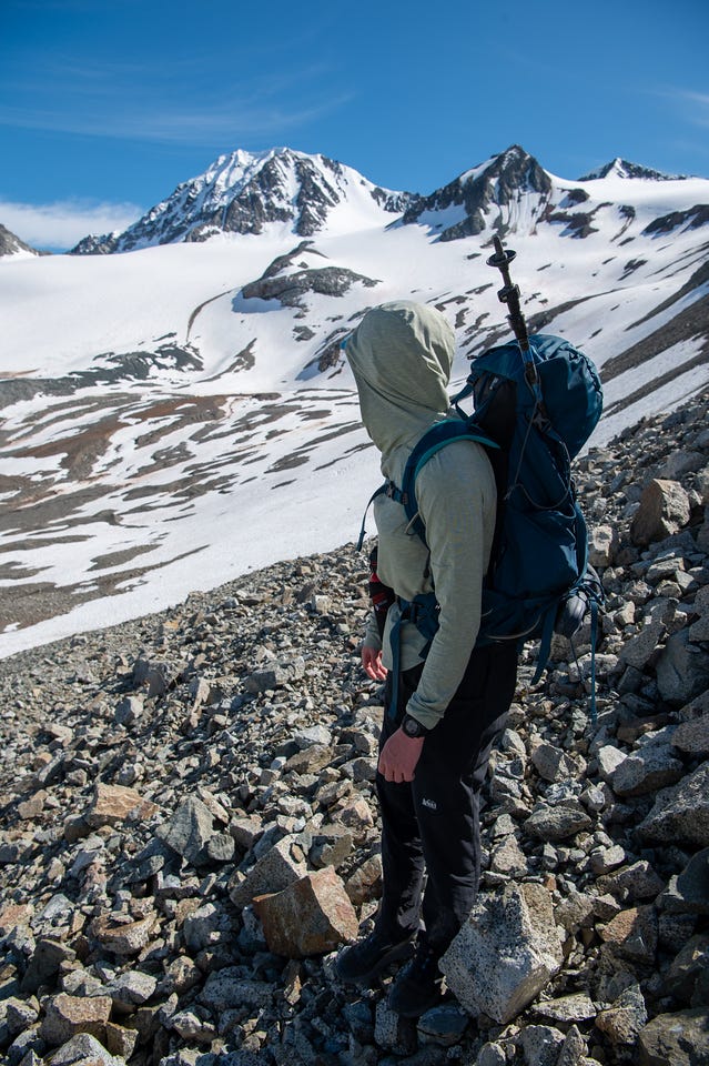 A hiker in a hood and large teal pack stands on a slope of loose rock, looking out across a snow-filled basin toward jagged snow-covered peaks. Old snow patches streak the slopes below under a clear blue sky.