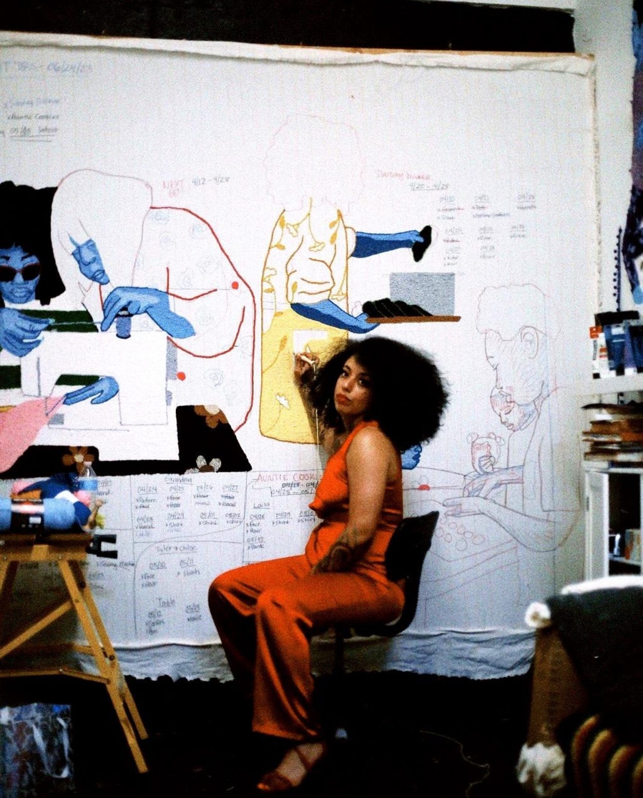 Honey Pierre seated in her studio, punch-needling a large textile painting with outlined blue figures on the wall.
