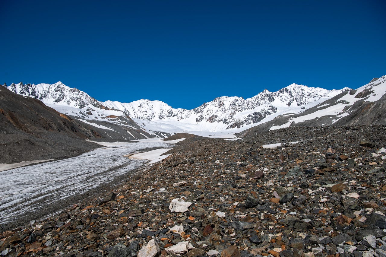 A wide view across a foreground of colorful glacial rubble — grey, rust-orange, and white cobbles — sloping down to a snow-covered glacier basin. Snow-plastered peaks ring the head of the valley, a broad tongue of dirty snow and ice curving down its center, all under a cloudless deep blue sky.