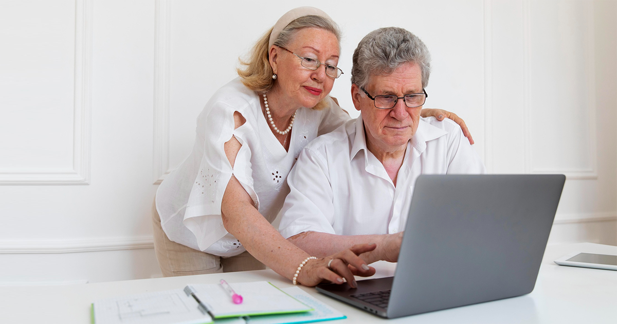 Um casal de idosos. O homem está sentado usando o notebook e a mulher está de pé mexendo no teclado. Os dois usam camisas claras e óculos. Ela usa colar, brinco e pulseira de pérolas, batom e sombra. Tem um acessório no cabelo, que é loiro e grisalho. Ele tem cabelo grisalho um pouco crespo. Um casal de idosos. O homem está sentado usando o notebook e a mulher está de pé mexendo no teclado. Os dois usam camisas claras e óculos. Ela usa colar, brinco e pulseira de pérolas, batom e sombra. Tem um acessório no cabelo, que é loiro e grisalho. Ele tem cabelo grisalho um pouco crespo.