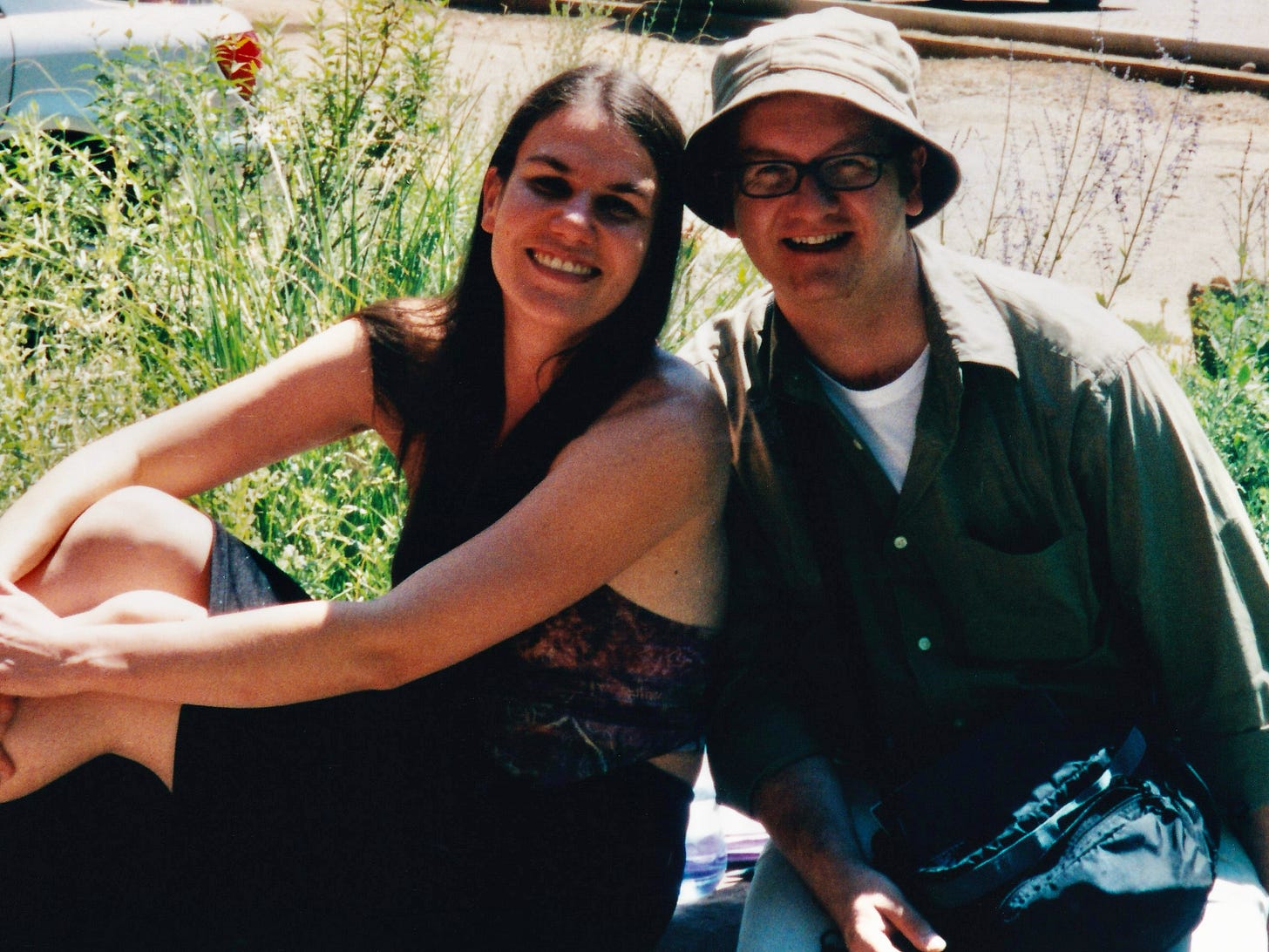 A woman and a man sit together outside. They are smiling at the camera. It is noon and very sunny. There are plants and a parked car behind them. 