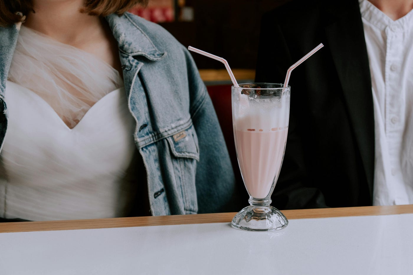 A close-up shot of a glass of milkshake with straws on a table. Seated on the left side, in front of the table, is a woman and a man — possibly her date — on the right. There are two straws in the glass of milkshake. A close-up shot of a glass of milkshake with straws on a table. Seated on the left side, in front of the table, is a woman and a man — possibly her date — on the right. There are two straws in the glass of milkshake.
