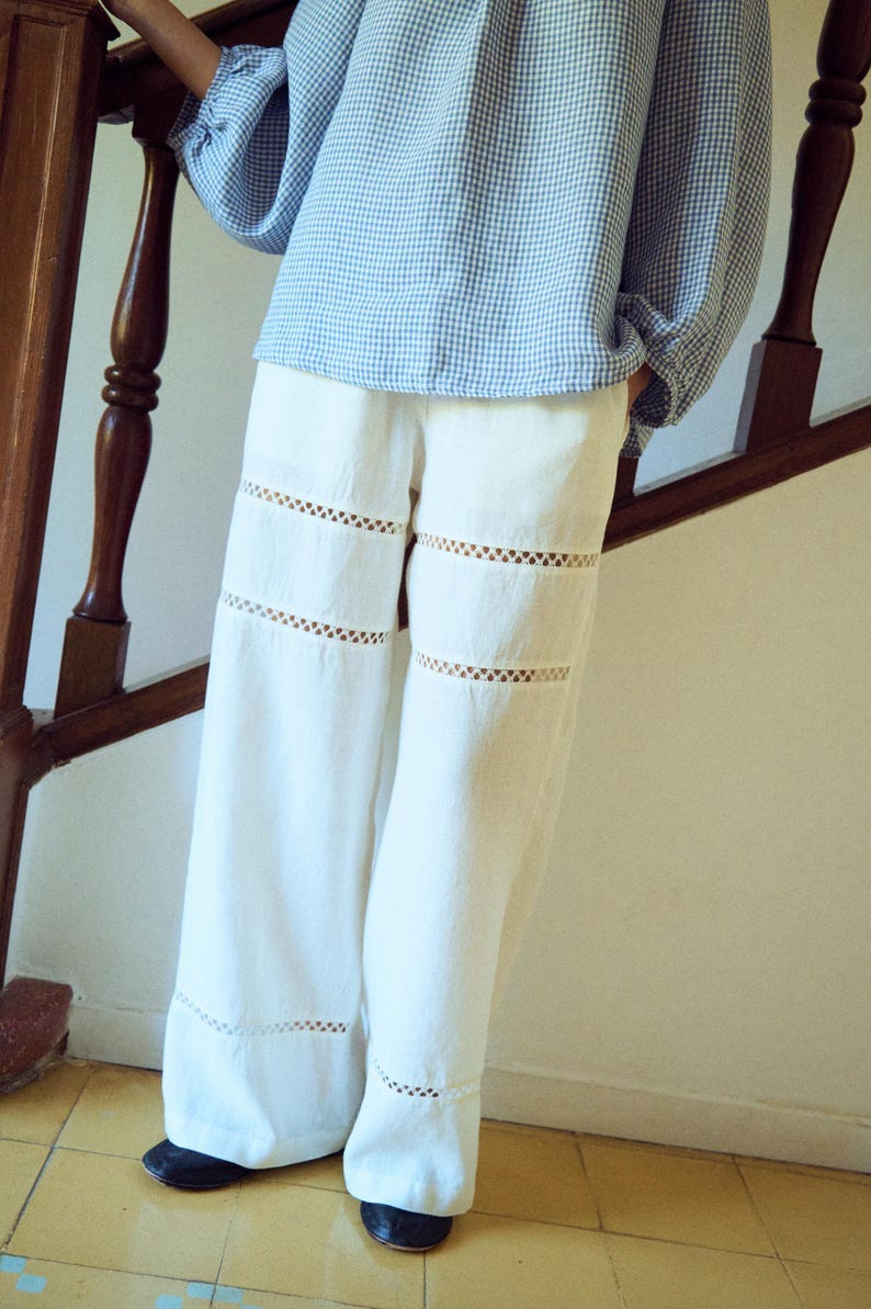 May include: White linen pants with decorative cut-out details. The pants have a wide leg design and are paired with a blue and white checkered blouse. The outfit is casual and suitable for warm weather.