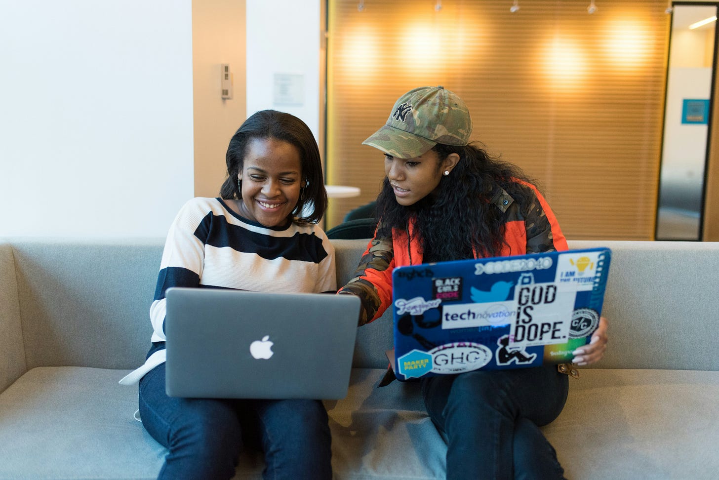 Two black female students, sitting together with laptops on a couch.