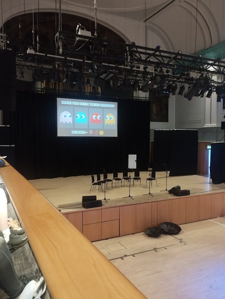 A theatre stage viewed from the balcony with seven chairs on the stage and pac-man images projected