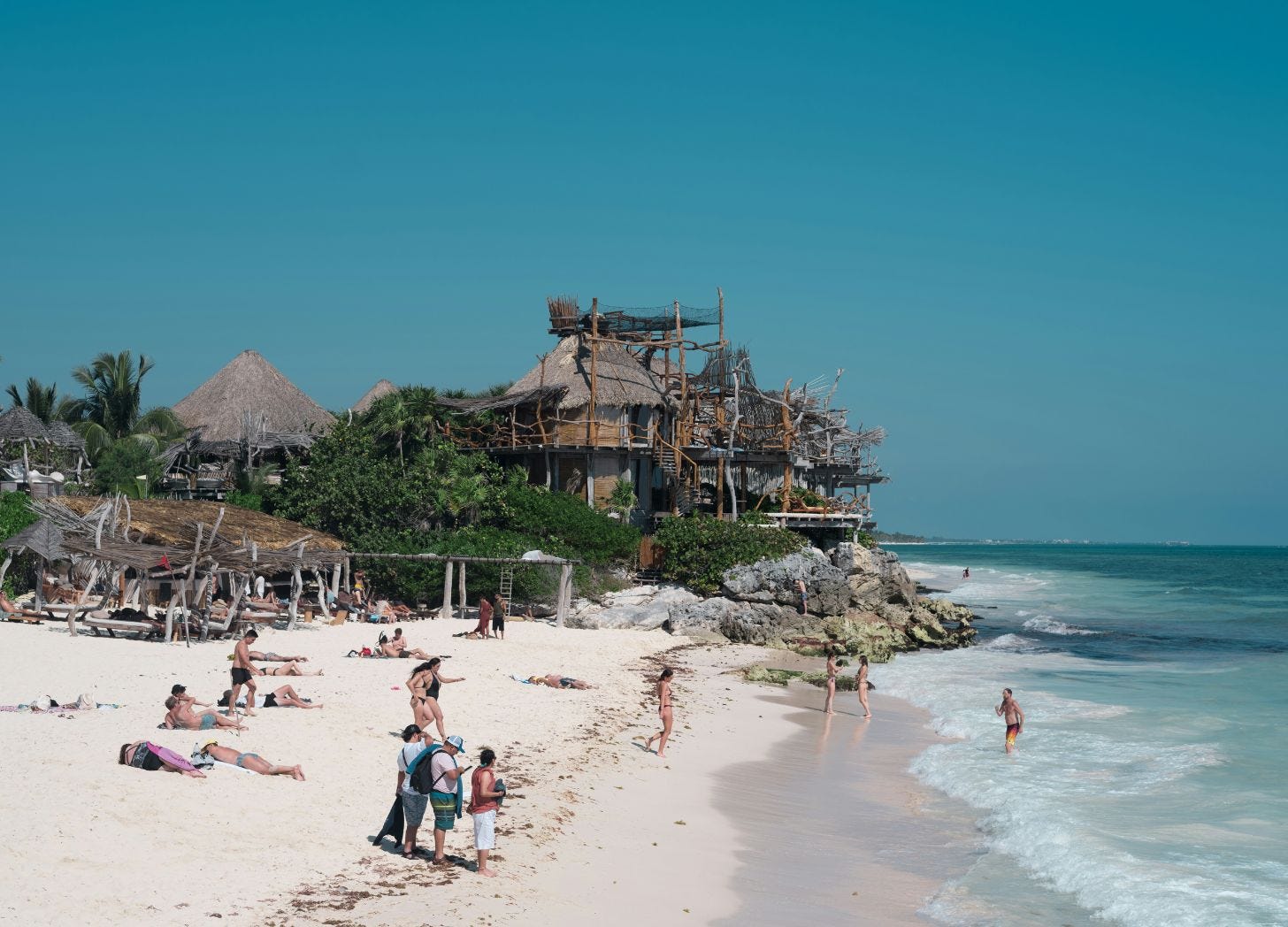 Crowded white-sand beach with people sunbathing and swimming, backed by rustic wooden and thatched buildings on a rocky shoreline. Tulum Mexico