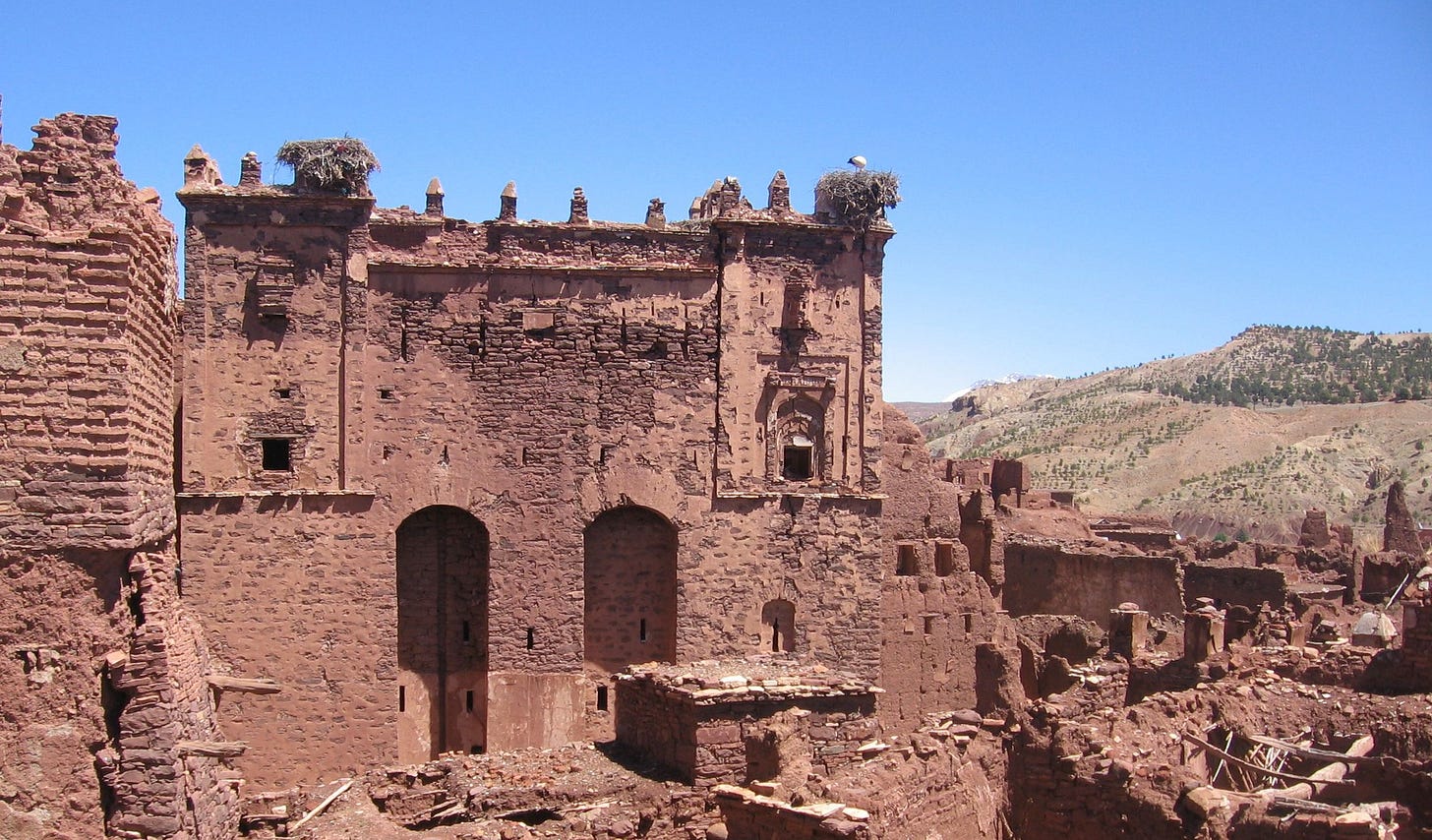 The ruins of a Moroccan fortress, built of red clay brick. Part has tumbled down. Beyond, enclosing the horizon, are scrubby desert hills.