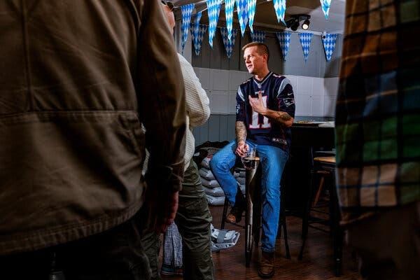 A man in a Patriots football jersey and bluejeans sits on a stool talking and holding a stein of beer. A man in a Patriots football jersey and bluejeans sits on a stool talking and holding a stein of beer.
