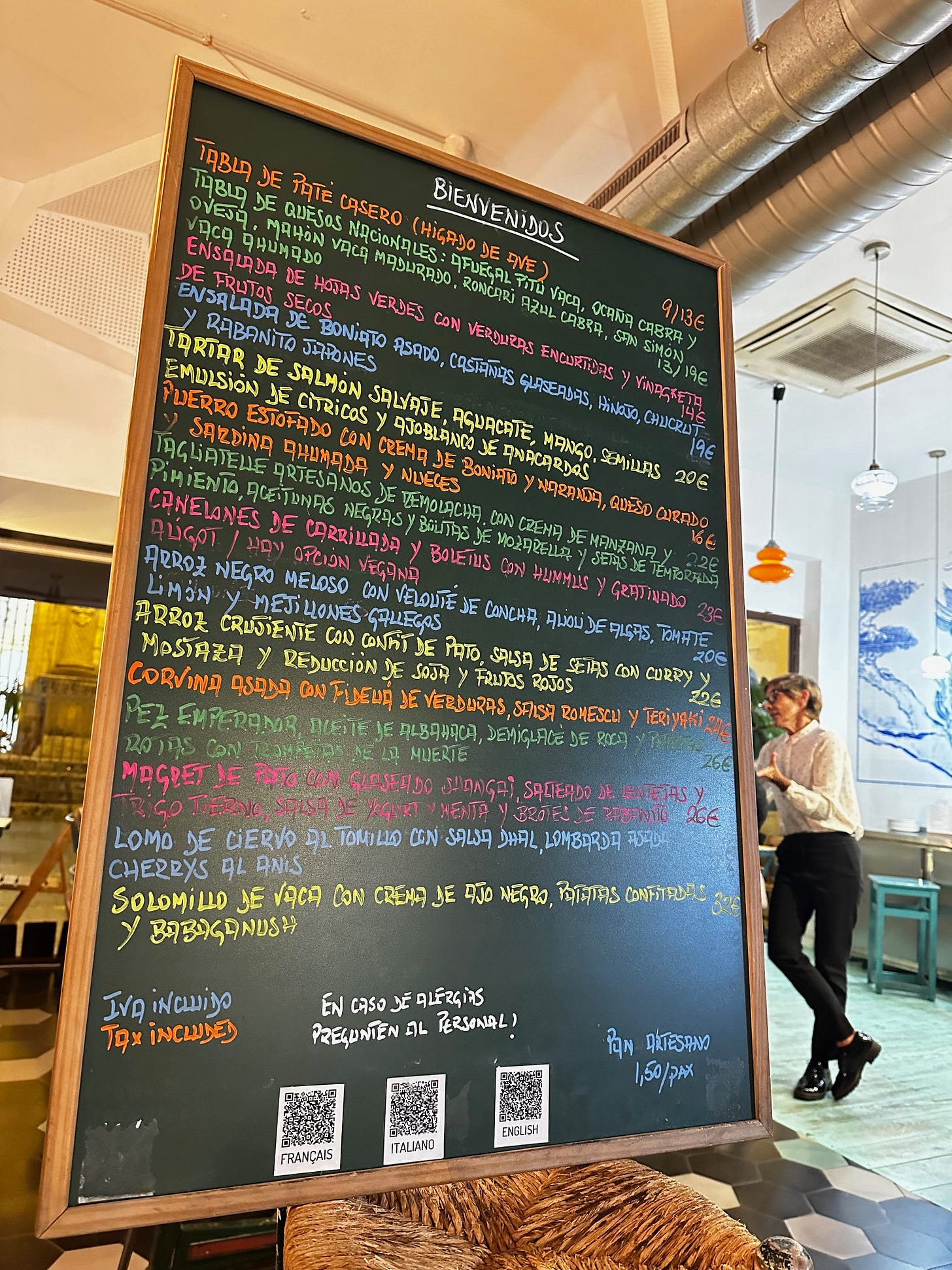 Chalkboard menu inside a restaurant in Sevilla, Spain. The board lists dishes written in colorful chalk with clear allergen notes and QR codes for French, Italian, and English translations. A server stands in the background, and the warm, casual interior creates a welcoming atmosphere. Chalkboard menu inside a restaurant in Sevilla, Spain. The board lists dishes written in colorful chalk with clear allergen notes and QR codes for French, Italian, and English translations. A server stands in the background, and the warm, casual interior creates a welcoming atmosphere.