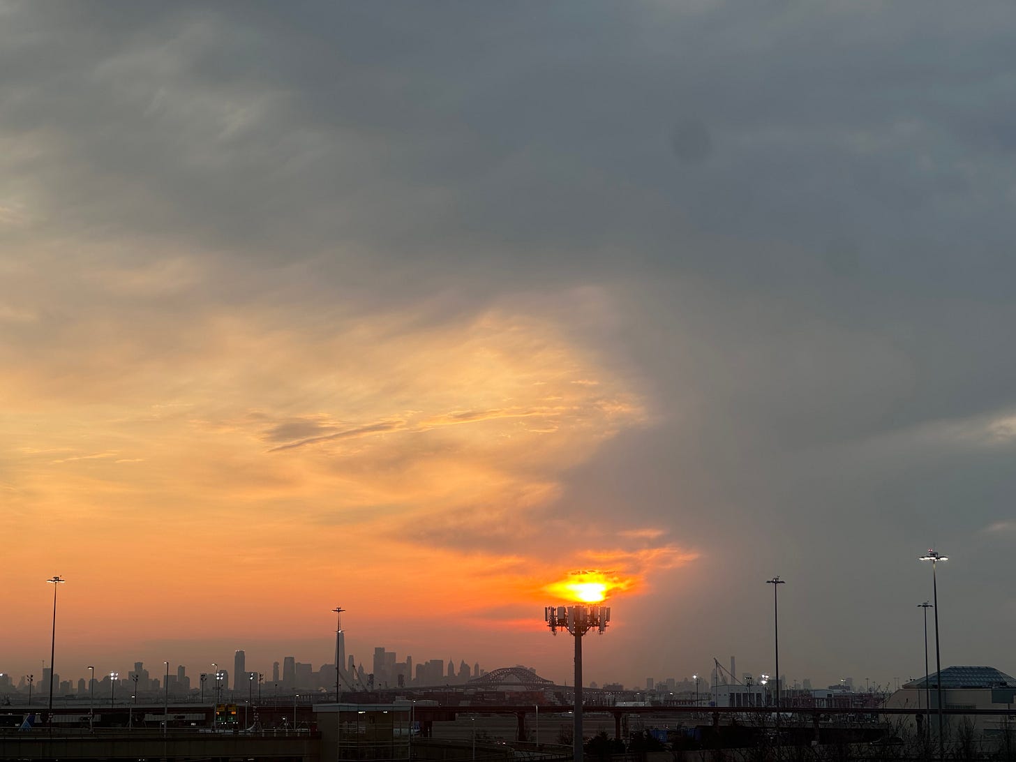 orange fireball of sun over New York City skyline, distant and small in background, with tall lights of extensive Newark Airport parking lots and runways in between