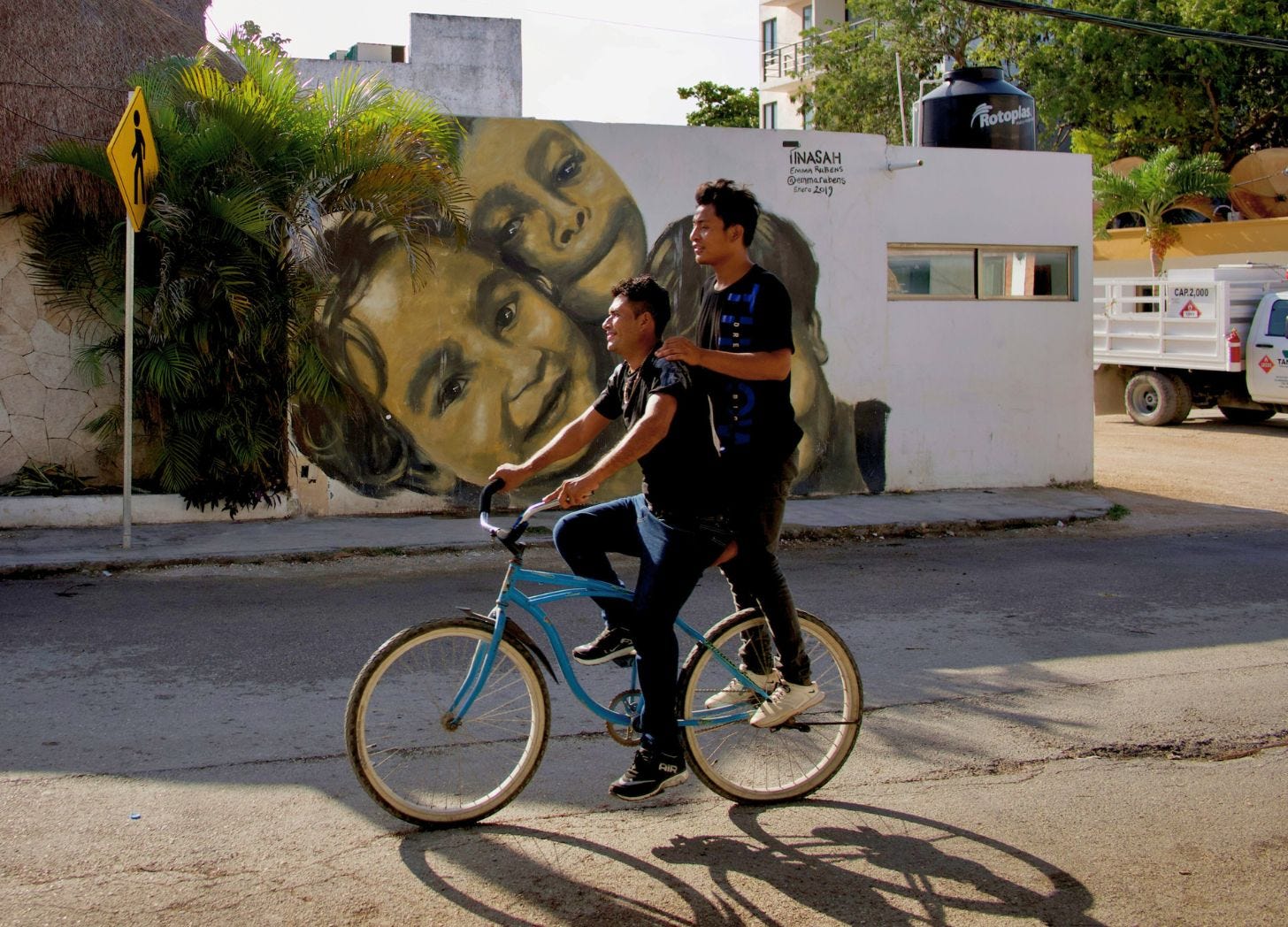 Two people riding a bicycle past a street mural depicting overlapping faces on a white wall in a small town street. Tulum Mexico