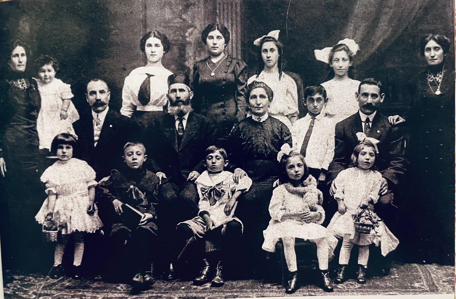 An elegant black and white family portrait of 17 people, adults and children in formal dress. At the center are the matriarch and patriarch, the man wearing a cylindrical head covering. The unmarried women and girls wear ribbons on their heads; the little girls hold small baskets, and one holds a stuffed bear. The expressions are serious and tired, save for the toddler in a woman's arms at the far left with a faint smile. 