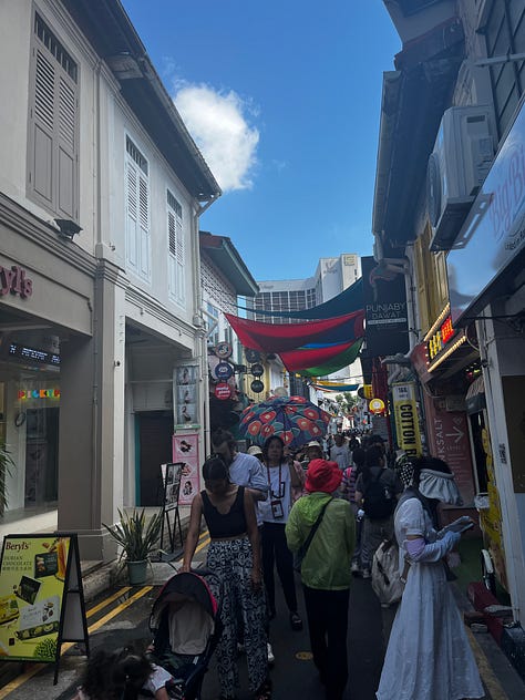 Top left photo is a green street sign that reads Haji Lane. Middle top photo is a white and blue shopping bag with Loka Made in print letters. Top right photo is a crowded street with shops on both sides of the lane. Bottom left photo is a street mural on Muscat Street. Bottom middle photo is of the Sultan Mosque. Bottom right photo is a gray mailbox and a stack of postcards being fed into the open slot. 
