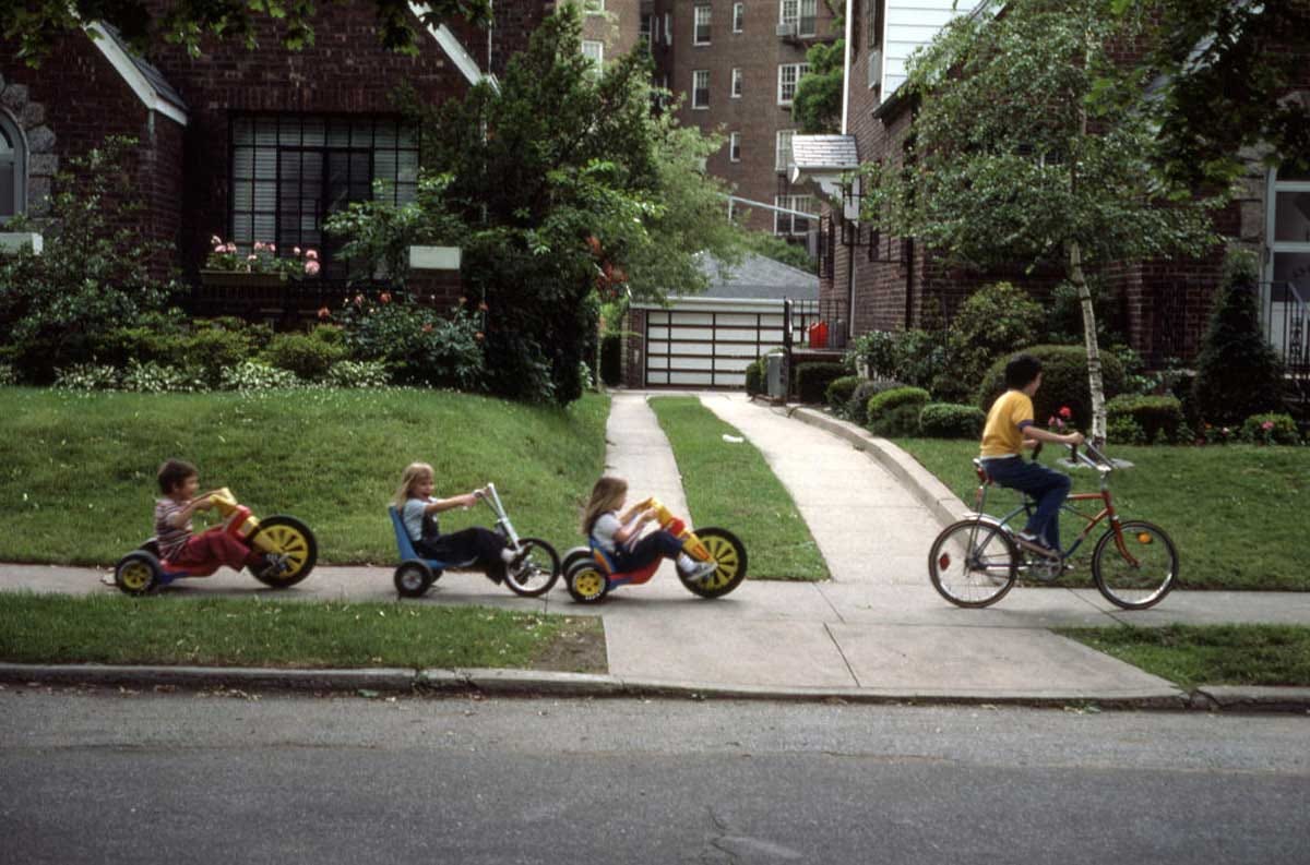 Three children ride tricycles in a row on a sidewalk, following an adult on a bicycle in a residential neighborhood with green lawns and brick houses. Three children ride tricycles in a row on a sidewalk, following an adult on a bicycle in a residential neighborhood with green lawns and brick houses.