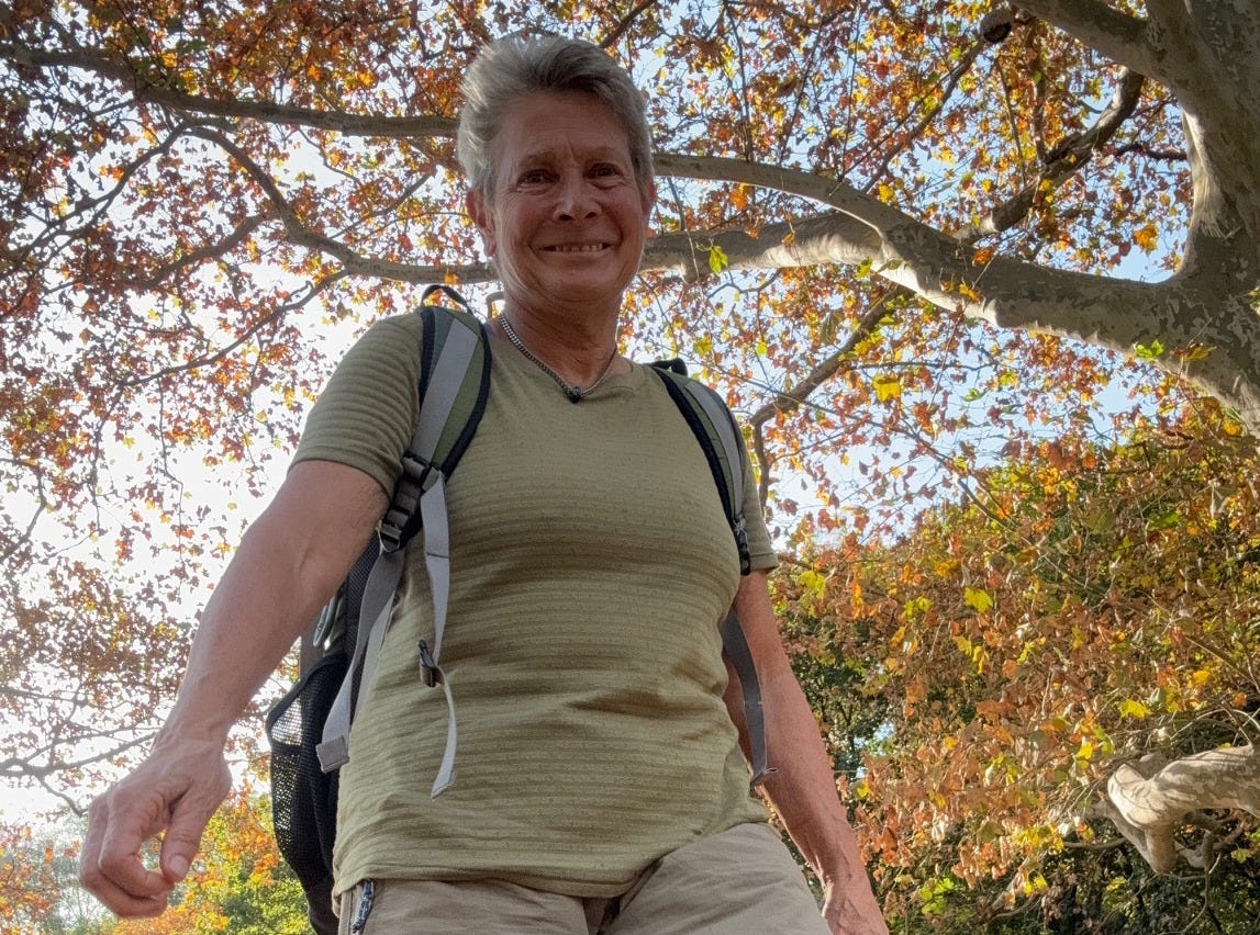 a woman with short, ash-colored hair looking down at a camera and smiling, against a fall scene. she is wearing a beige textured t-shirt with horizontal stripes and a backpack