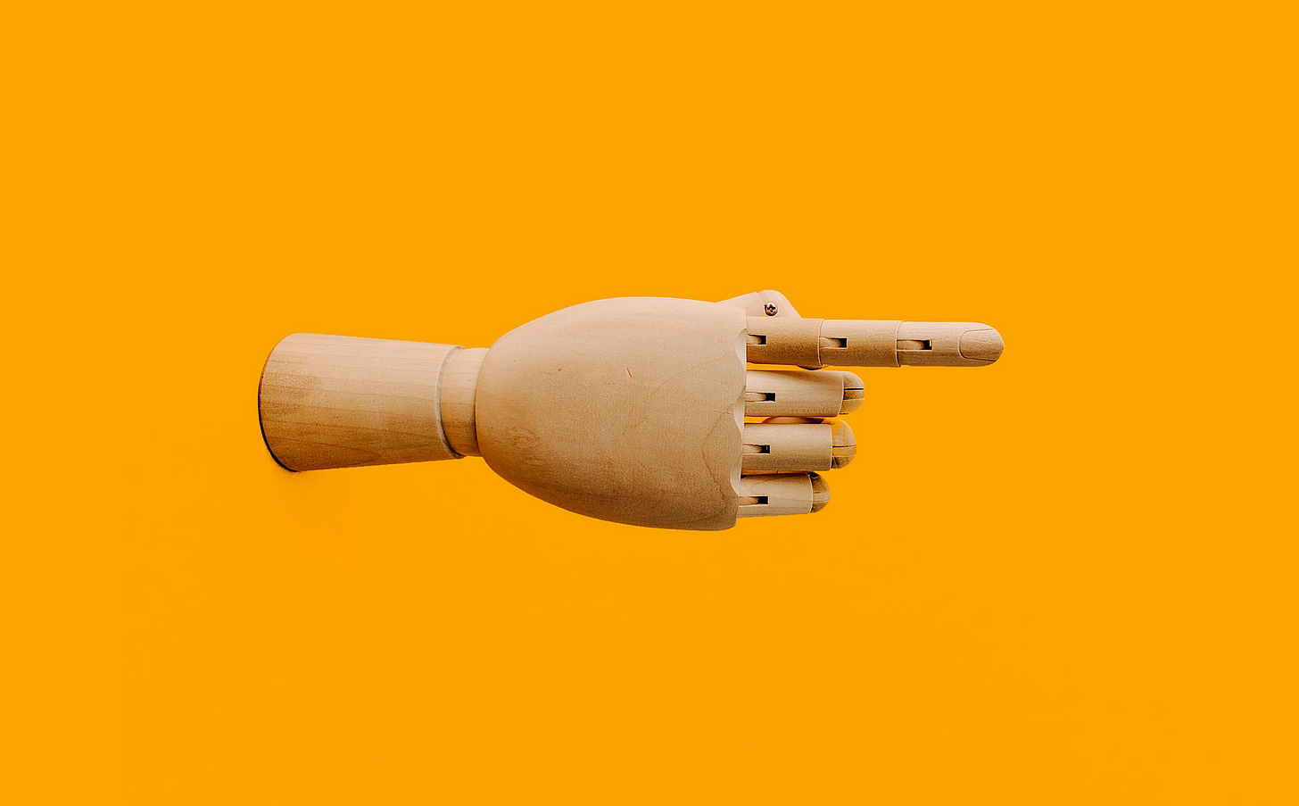 A wooden hand arranged so that its index finger is pointing. The image has been rotated so that the finger is pointing to the right. The image consists of just the hand against a solid orange background. A wooden hand arranged so that its index finger is pointing. The image has been rotated so that the finger is pointing to the right. The image consists of just the hand against a solid orange background.