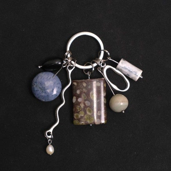 May include: A silver keychain with various decorative charms. The charms include a blue sphere, a rectangular stone with brown and green spots, a small white pearl, and other geometric shapes. The background is solid black.