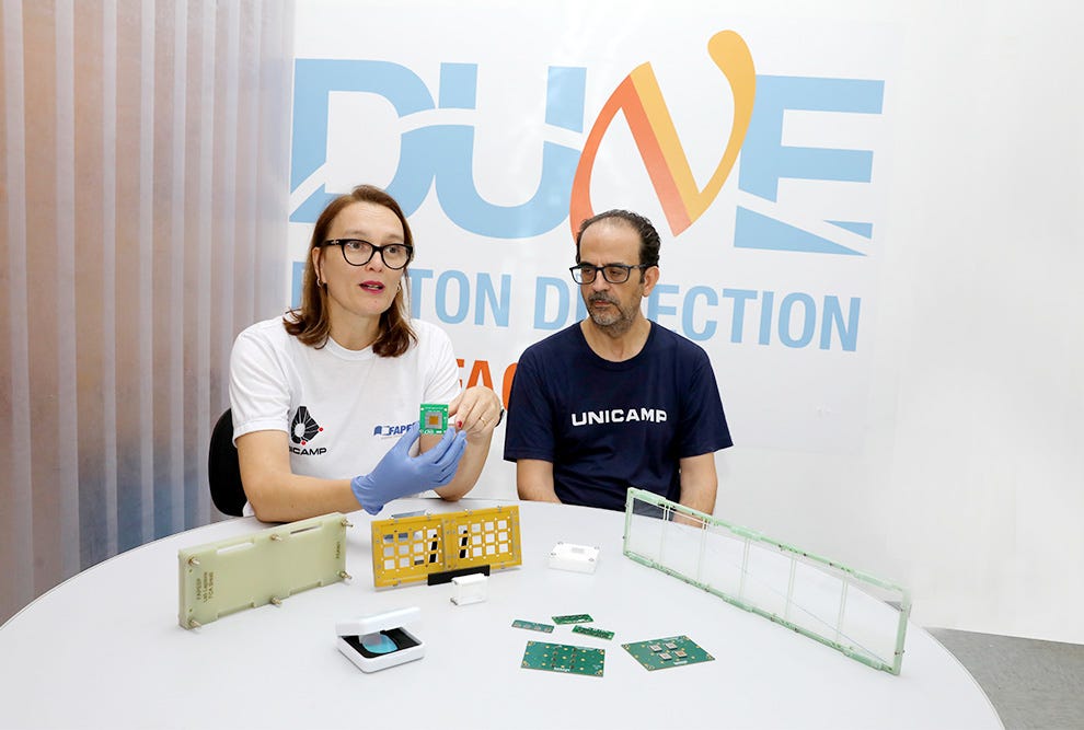 Duas pessoas sentadas à uma mesa branca em frente a um painel com logo "DUNE" e texto "TON DIRECTION" em azul e laranja, sendo que a pessoa à esquerda veste camiseta branca com logotipo, óculos e luvas azuis enquanto segura um componente eletrônico verde, e a pessoa à direita veste camiseta azul escuro com texto "UNICAMB" e óculos, ambas observando diversos componentes eletrônicos, placas de circuito verde, um dispositivo branco e uma caixa amarela organizadora dispostos sobre a superfície da mesa. Duas pessoas sentadas à uma mesa branca em frente a um painel com logo "DUNE" e texto "TON DIRECTION" em azul e laranja, sendo que a pessoa à esquerda veste camiseta branca com logotipo, óculos e luvas azuis enquanto segura um componente eletrônico verde, e a pessoa à direita veste camiseta azul escuro com texto "UNICAMB" e óculos, ambas observando diversos componentes eletrônicos, placas de circuito verde, um dispositivo branco e uma caixa amarela organizadora dispostos sobre a superfície da mesa.