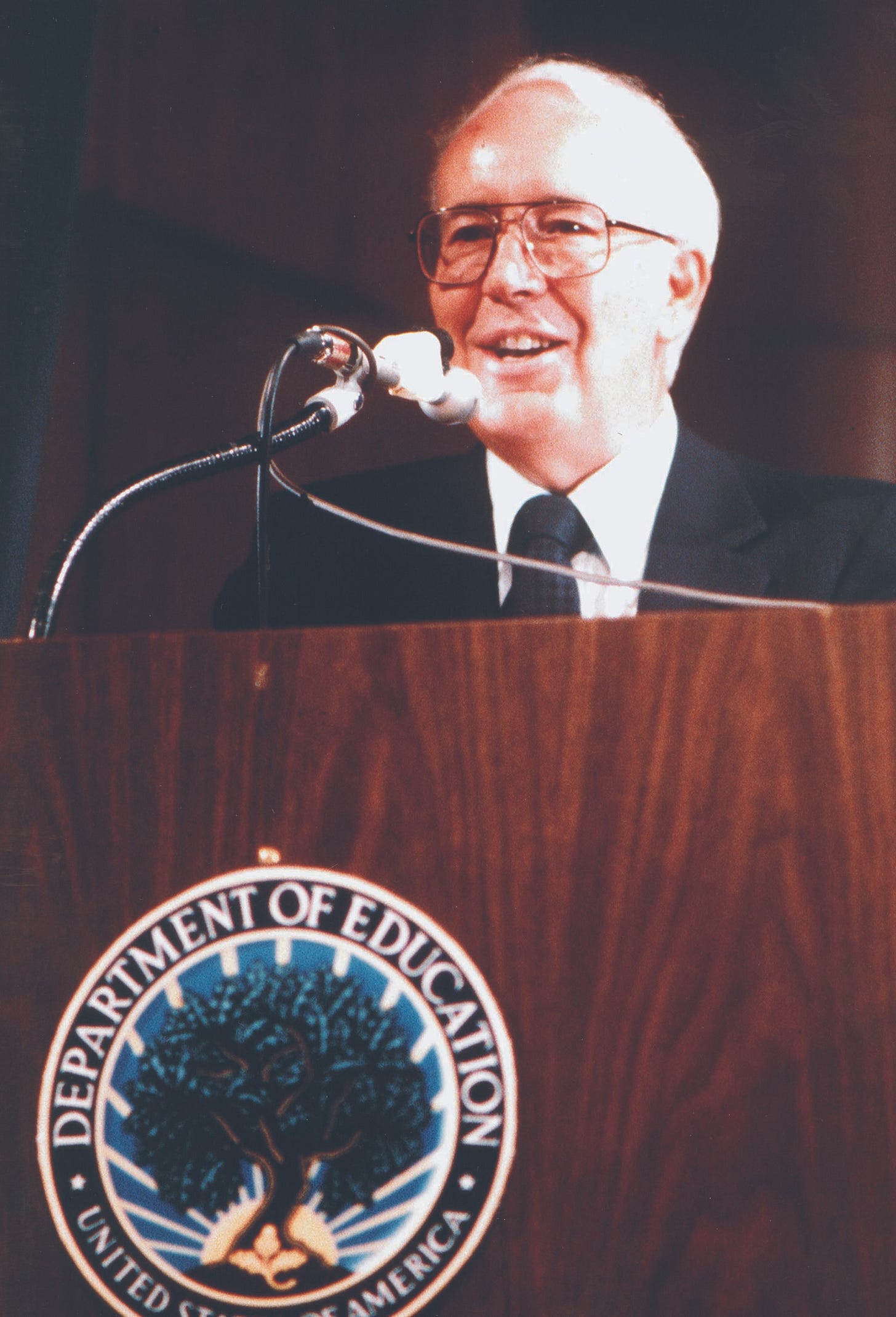 Terrel Bell, a white man with glasses and thinning white hair, speaks at a podium which bears the seal of the Department of Education