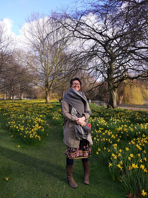 Three photos of me in St. James' Park in February 2020. I am standing surrounded by daffodils, and whilst I am rugged up in my grey coat and blanket scarf, a floral skirt peeks out from underneath and I am clearly embracing the new season. The background includes three big tree still in Winter mode but a small patch of blue sky is peeking through the clouds. Photos taken by S-