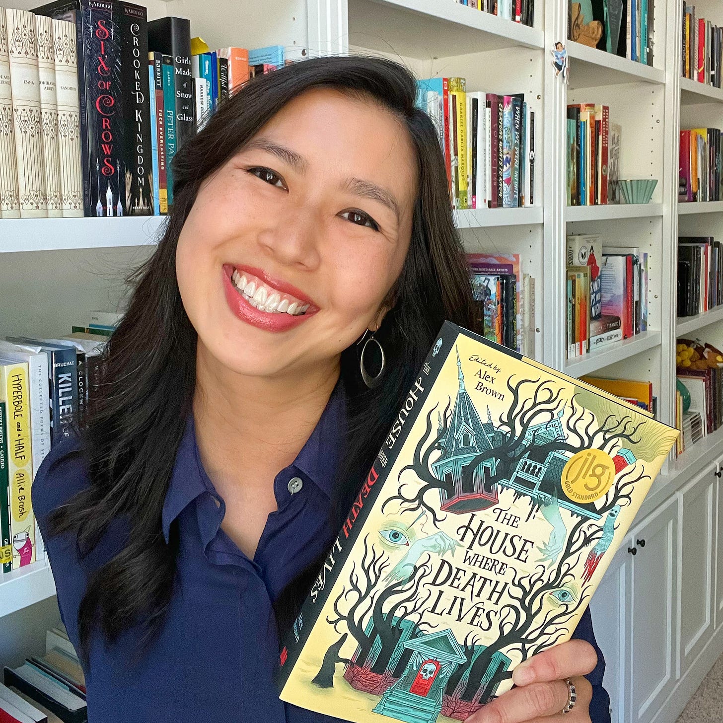 Traci Chee in a navy blouse, smiling and holding up a hardcover of THE HOUSE WHERE DEATH LIVES in front of some built-in bookshelves
