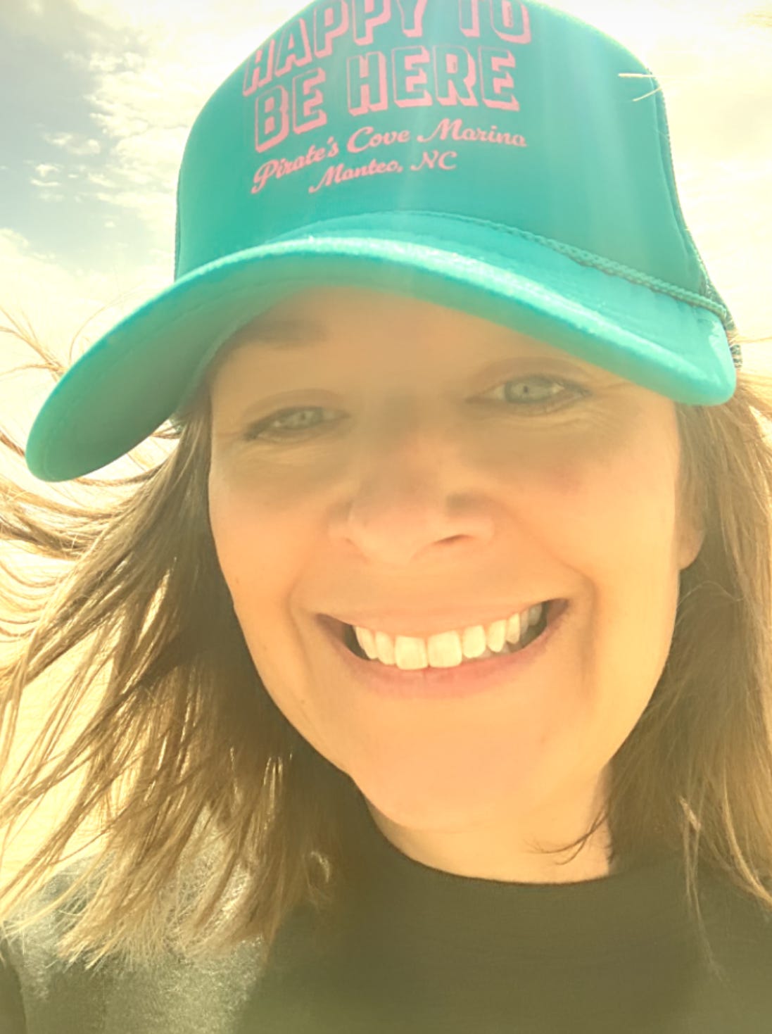 photo of me at the beach with wind in my hair, a toothy grin, and a hat that reads "happy to be here"