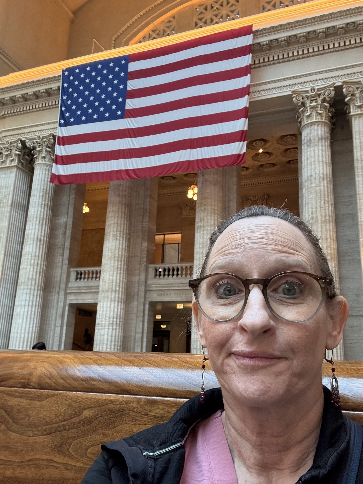 A woman looks quizzically at the camera while posing for a selfie in front of marble pillars and a large US flag in Chicago's Union Station