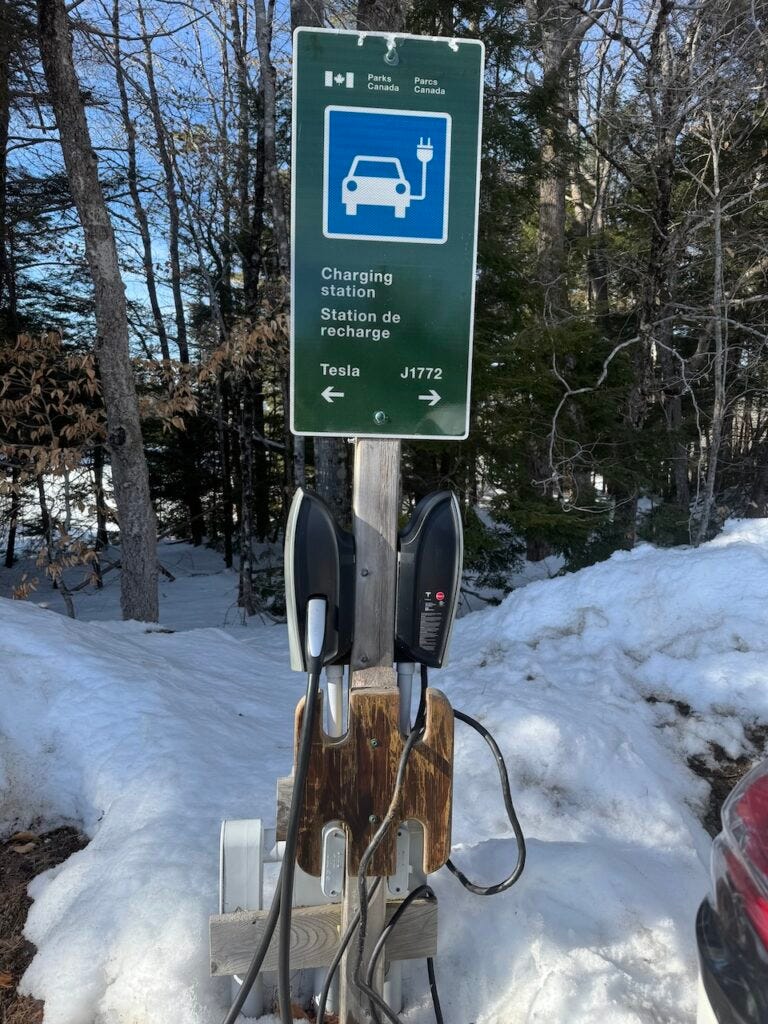 A Parks Canada/Parcs Canada sign marks an EV charging station, with two wall?mounted chargers on a wooden post amid snowy forest parking area.