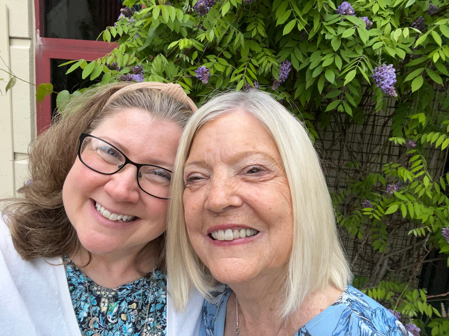 Close-up selfie of Patty and Sherry, heads tilted together, smiling, with flowers and greenery in the background.