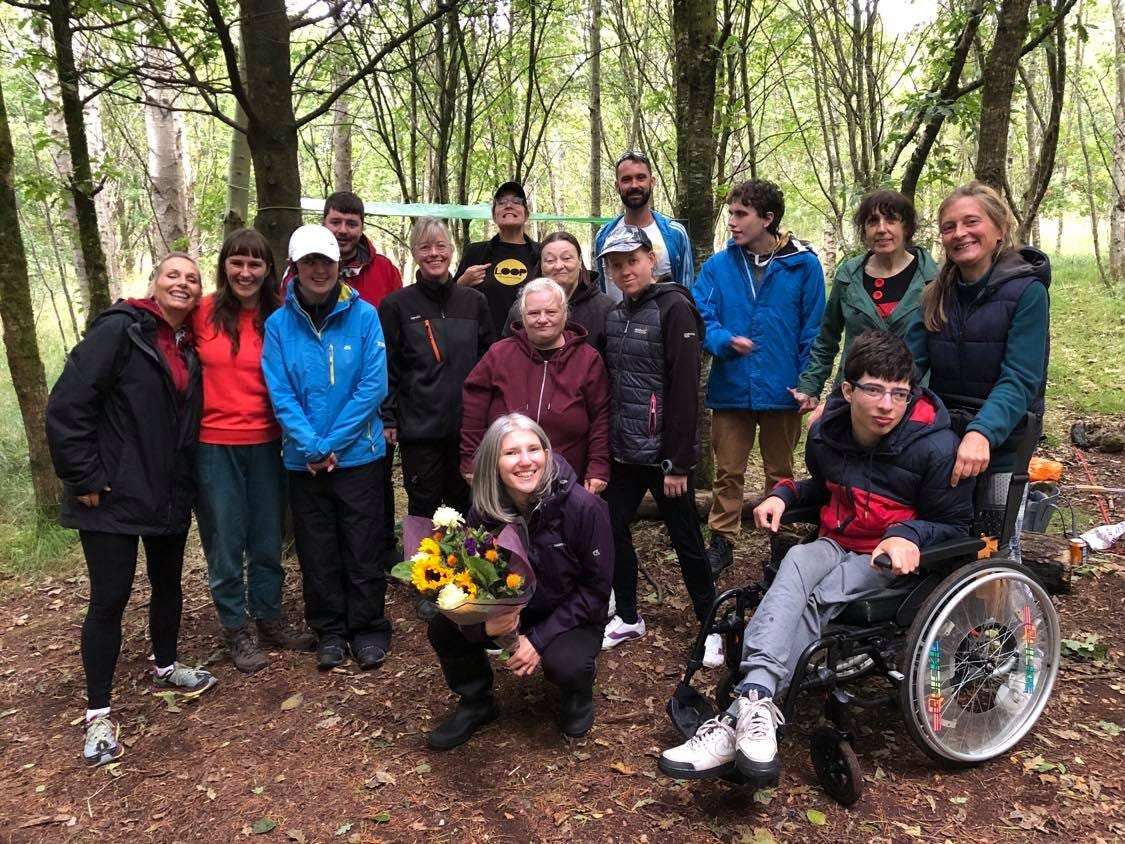 A group of people of different ages and genders, all white, gather together in a forest. There is a young person in a wheelchair at the forefront on the right side and on the left, a woman kneeling and holding a bunch of flowers. They are all dressed in outside wear and some wear black and yellow Loop t-shirts.