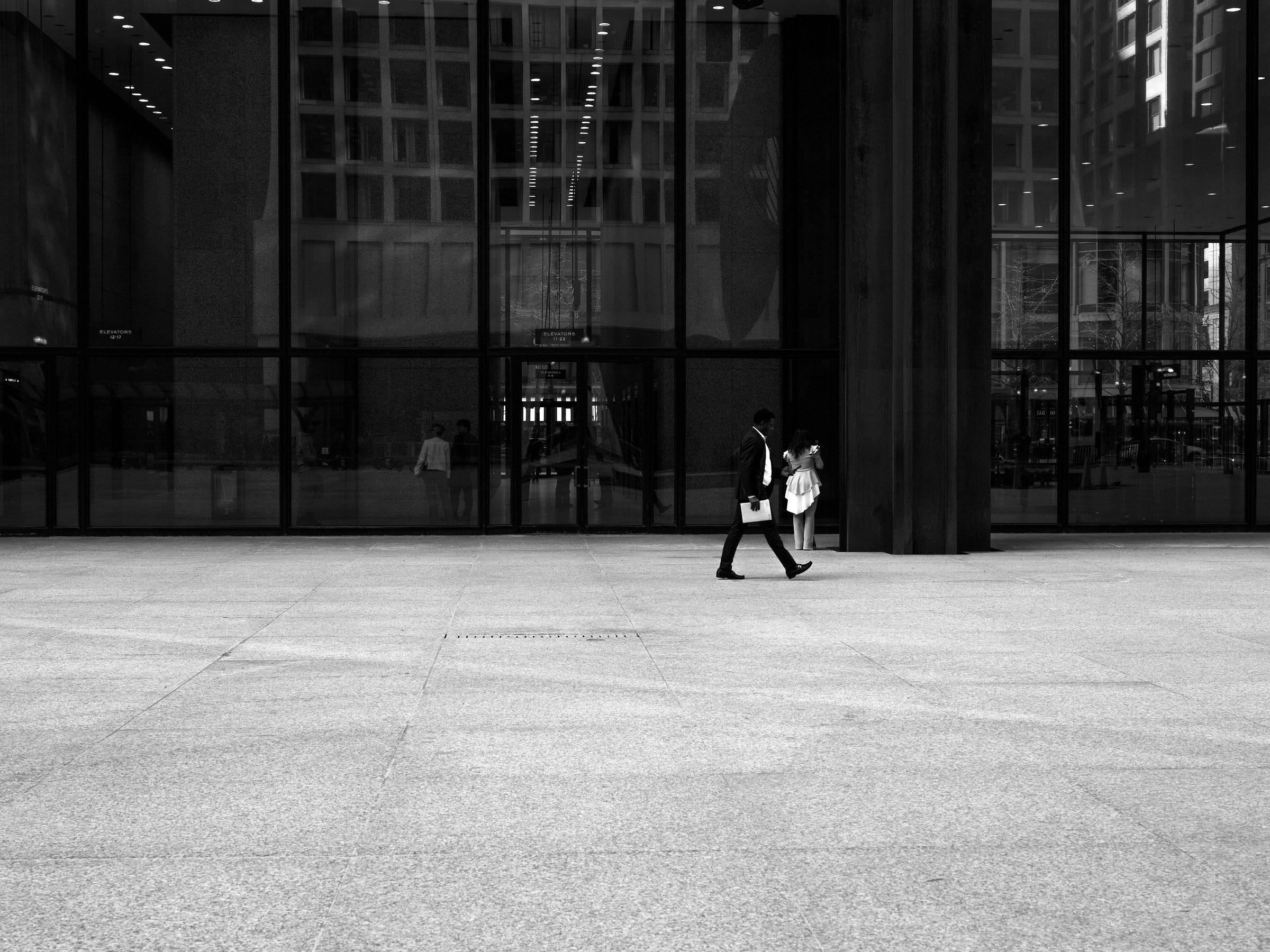 Black and white photograph of a businessman walking across a vast, open plaza in front of a modern glass building. He carries a folder and looks ahead with purpose. In the background, a young girl in a light dress leans against a column, seemingly lost in thought, while reflections of other pedestrians appear in the building’s windows. Black and white photograph of a businessman walking across a vast, open plaza in front of a modern glass building. He carries a folder and looks ahead with purpose. In the background, a young girl in a light dress leans against a column, seemingly lost in thought, while reflections of other pedestrians appear in the building’s windows.