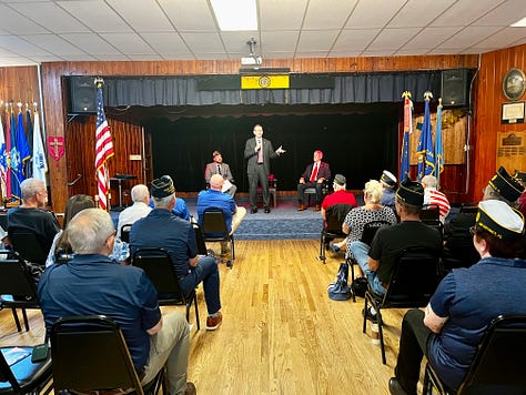 The image shows a diverse group of people seated in rows of chairs in a wooden-paneled hall, attentively facing forward. Many attendees are older adults, and some are wearing uniforms or hats indicating military or veteran status. The room is decorated with framed photos, memorabilia, and a disco ball hanging from the ceiling. There are several empty chairs with brochures or program booklets placed on them in the foreground. The atmosphere suggests a formal gathering or community event, likely related to veterans or a commemorative occasion. No specific brands, landmarks, or recognizable public figures are visible.
