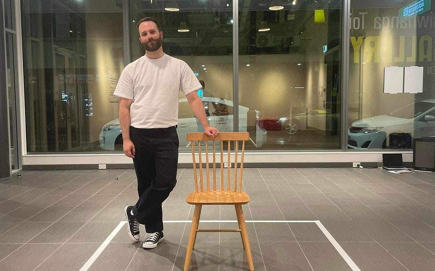 A photo of me standing in Te Auaha Gallery after a performance of Access, leaning on a wooden chair in a taped-off square on the floor. I’m wearing a white t-shirt, black pants, and Chucks, with a tired expression, in front of the large gallery windows.